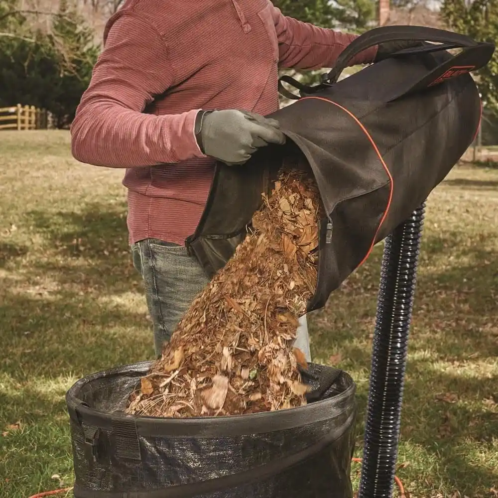 Gloved hand pouring mulched leaves from the BLACK+DECKER vacuum collection bag into a yard waste bin