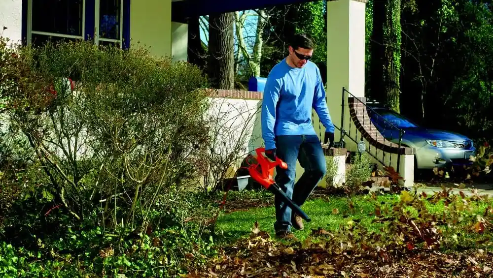 Man in a blue shirt blowing fallen leaves across a lawn in front of a house with a corded blower