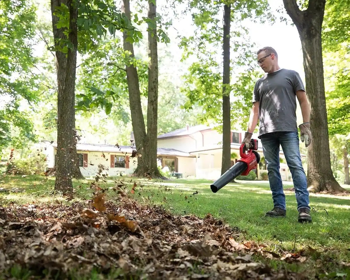 Man blowing a pile of leaves across a lawn between tall trees with the CRAFTSMAN V20 blower