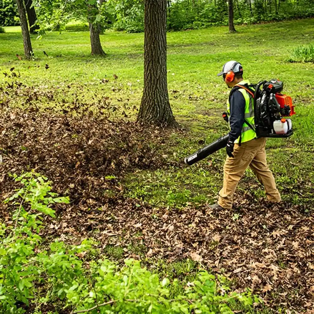 Landscaper in a safety vest and ear protection blowing leaves across a park lawn with a backpack blower