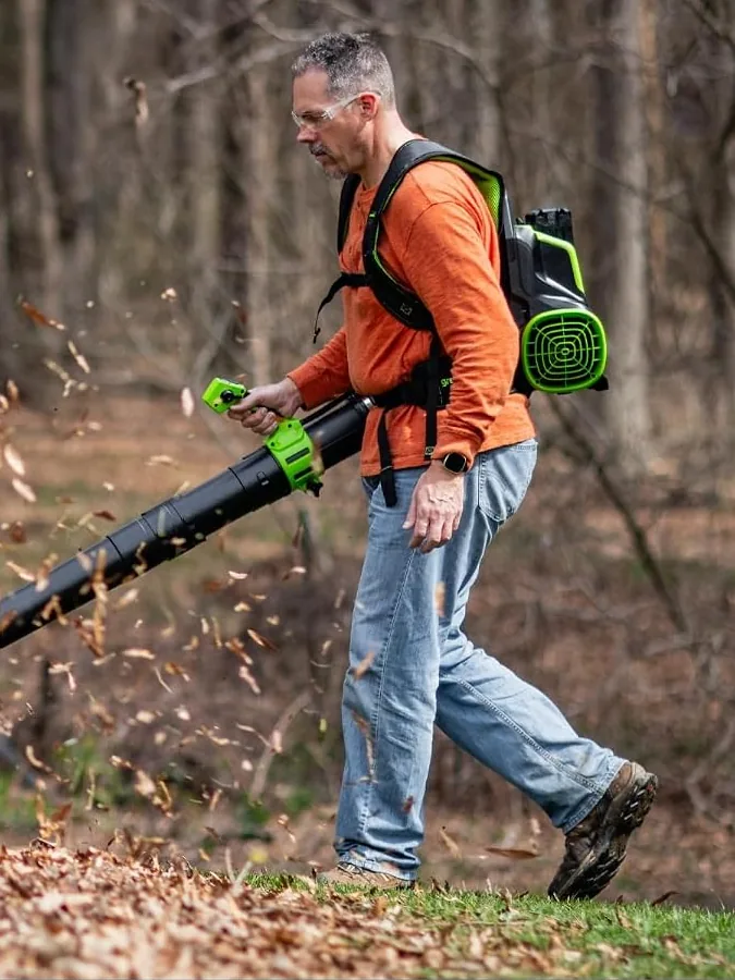Man in an orange shirt walking across leaf litter with the Greenworks 80V backpack blower on his back