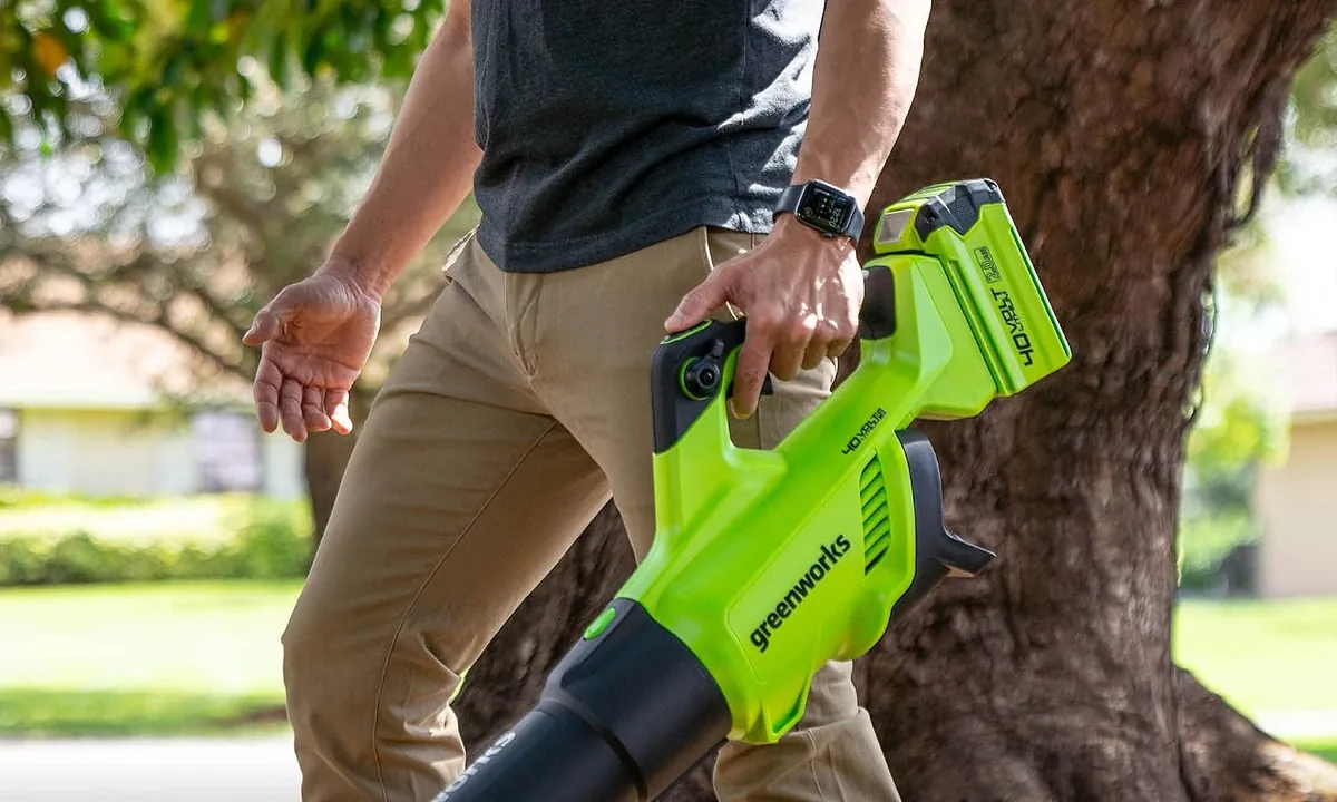 Man in khaki trousers carrying the Greenworks 40V blower with a battery fitted, walking past a tree