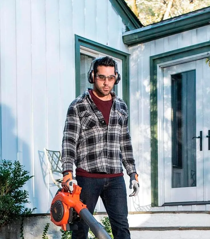 Man in ear protection and safety glasses carrying a Husqvarna 125B handheld blower on a stone patio