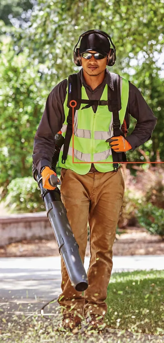 Worker in a safety vest and ear muffs walking a lawn with a Husqvarna backpack blower