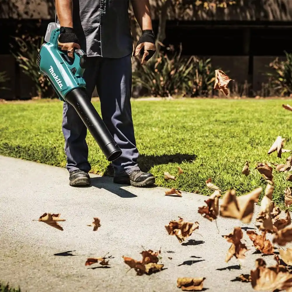 Worker in gloves holding the Makita XBU03Z blower beside a lawn while leaves blow across the path