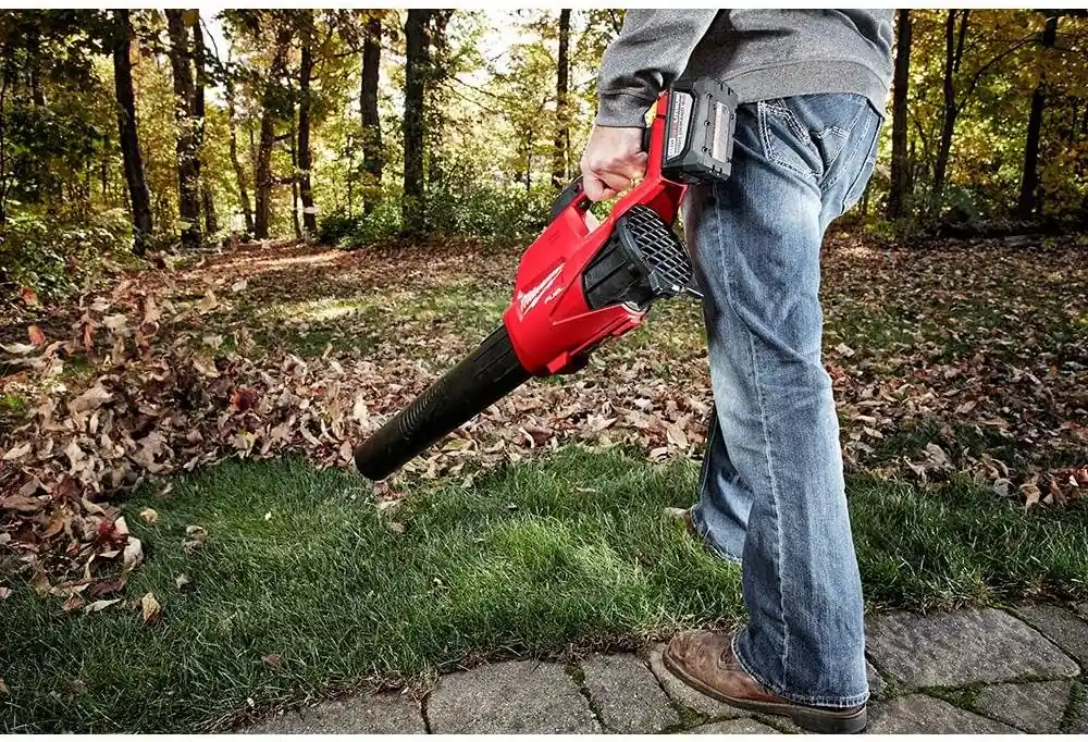 Man in jeans carrying the Milwaukee M18 FUEL blower over a lawn covered with autumn leaves